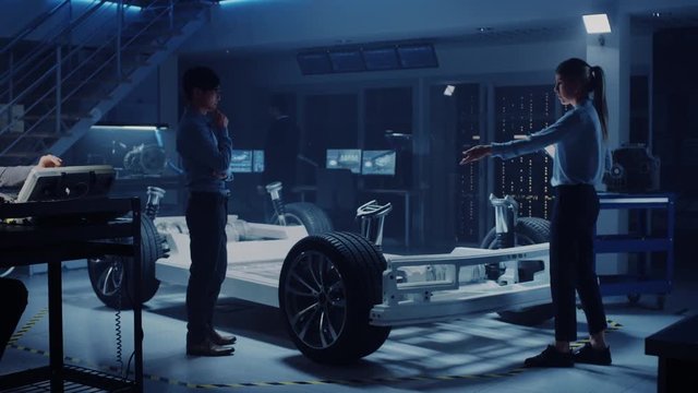 Automotive Design Engineer and Scientist with Tablet Discuss the Electric Car Chassis Prototype. In Innovation Laboratory Facility Concept Vehicle Frame Includes Wheels, Suspension, Engine and Battery