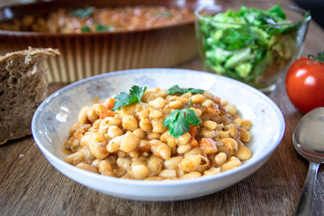 Stewed and baked white beans with spices in a plate and lettuce and tomato salads with homemade bread on wooden table