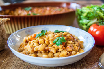 Stewed and baked white beans with spices in a plate and lettuce and tomato salads with homemade bread on wooden table