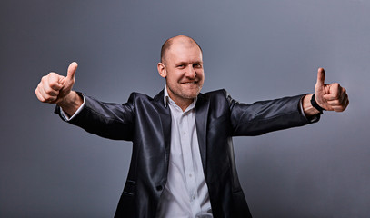 Fun comic bald business man in black suit showing the finger success thumb up sign on grey...