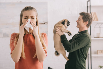selective focus of blonde girl allergic to dog sneezing in white tissue near cheerful man holding pug