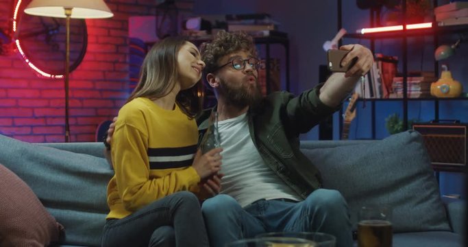 Young good looking together and happy Caucasian couple sitting on the couch in the cozy room with drinks and posing to the smartphone camera while taking selfie photos. At night.