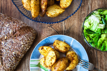 Baked young potatoes on a plate, homemade whole grain bread and lettuce salad on vintage wooden table