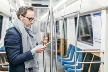 Naklejka na ściany i meble Young man with glasses and wireless headphones rides in the subway train. He is wearing a blue coat and a gray scarf. In his hands he holds mobile phone. Digital dependence, social networks addiction.