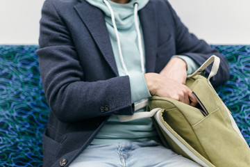 Close up photo of sitting hipster in blue coat and a turquoise hoodie who pulls something out from green backpack. A man pulls out a book/newspaper to read while riding the subway. Urban life concept.