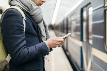 Close up portrait of attractive man in blue coat who looks at his phone and writes a text message. Selective focus. Social networks addiction. Waiting for the train at subway station platform.