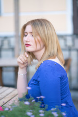 charming caucasian blonde girl in a blue dress sitting at a table in a city cafe alone and waiting