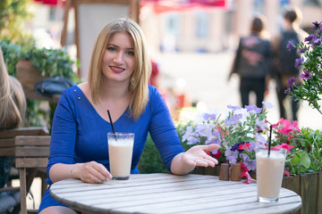 a charming caucasian girl in a blue dress sitting at a wooden table in a street cafe and invites you to sit down and drink coffee on this Sunny summer day