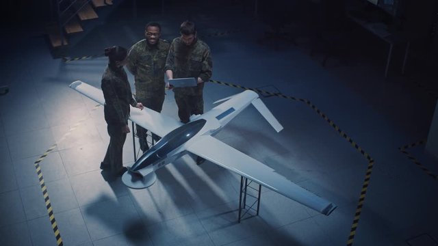 Army Aerospace Engineers Work On Unmanned Aerial Vehicle / Drone. Uniformed Aviation Experts Talk, Using Laptop. Industrial Facility with Aircraft for Performing Surveillance, Warfare Tactics, Attack