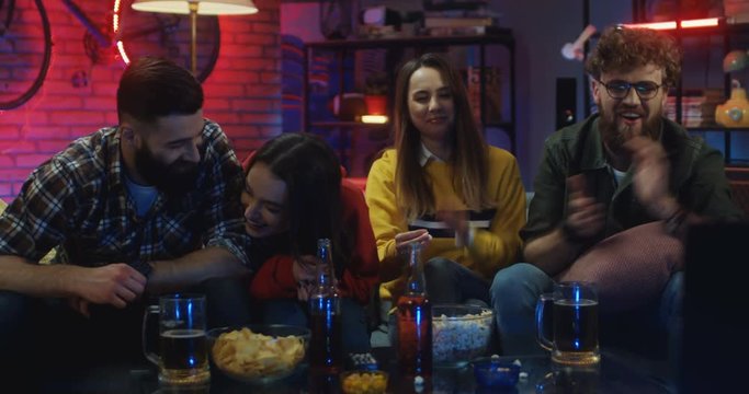 portrait shot of the Caucasian young cheerful friends sitting on the couch and laughing while watching comedy movie or show on TV in the living room at night.