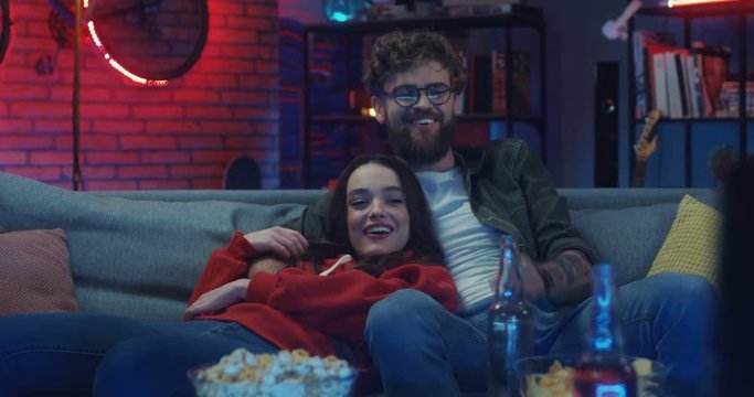 Pretty Caucasian young girl and handsome guy resting on the sofa at night, smiling and watching movie on TV.