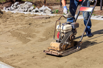 Worker starts a gasoline compactor for ramming sandy soil for laying paving slabs.