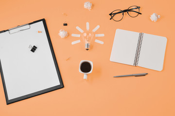 Flat lay financial planning brainstorming messy table top image with blank clip board, office supplies, pen, notepad, eyeglasses, cup of coffee, light bulb on orange background.