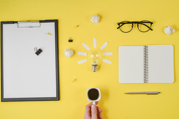 Flat lay brainstorming table top view. Blank clip board, office supplies, pen, notepad, eyeglasses, cup of coffee in hand, light bulb on yellow background. Concept new idea