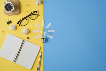 Flat lay financial planning brainstorming messy table top image with blank clip board, office supplies, pen, notepad, eyeglasses, coffee cup, light bulb on yellow and blue background.