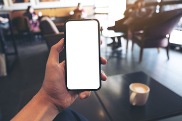 Mockup image of a hand holding and showing black mobile phone with blank white screen in cafe