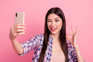 Close-up portrait of her she nice attractive cute charming lovely lovable cheerful straight-haired lady in checked shirt holding in hand cell making selfie showing ok-sign isolated on pink background
