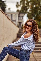 Young happy woman with long brown hair in sunglasses, blue jeans, blue shirt walks through the old town on a cloudy summer day.