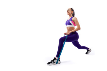 Fototapeta na wymiar A dark-haired woman coach in a sporty purple short top and gym leggings makes lunges with sport fitness rubber bands on a white isolated background in studio