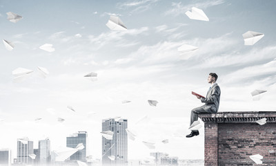 Man on roof edge reading book and cityscape at background