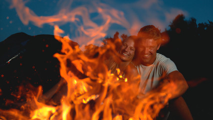 CLOSE UP: Excited woman laughs while her boyfriend pokes the campfire with stick
