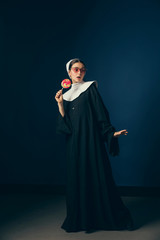 Being stylish every day. Medieval young woman as a nun in vintage clothing, white mutch and red sunglasses eating sweets and standing on dark blue background. Concept of comparison of eras.