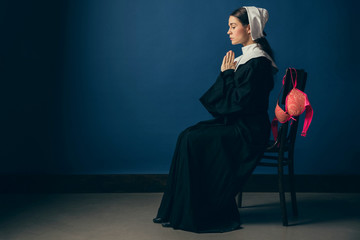 Memories of youth or crazy weekend. Medieval young woman as a nun in vintage clothing and white mutch sitting on the chair with bright pink bra on blue background. Concept of comparison of eras.