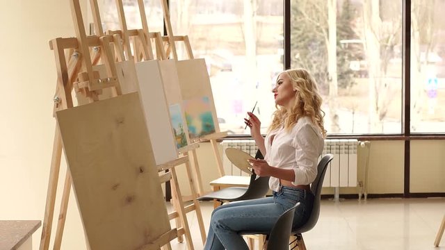 A young sexy girl in a white shirt draws on canvas in the studio for drawing among a variety of paintings, against a large window. Art.