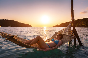 Woman relaxing at the beach in a hammock above sea water, warm sunny summer evening at vacation holidays hotel, happy moment at sunset