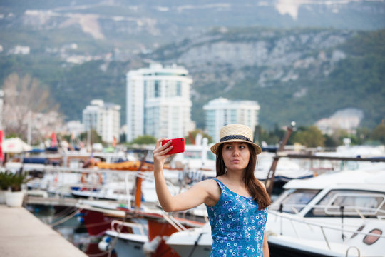 Young travelling woman in hat taking selfie photos over summer sunny mediterranean city with smartphone camera. Brunette girl making photography on summer vacation