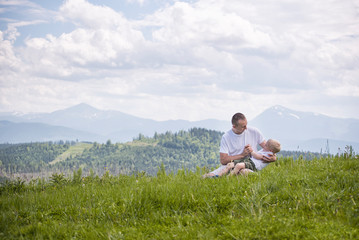 Naklejka na ściany i meble Happy father with his little son on his knees sits against a background of green forest, mountains and sky with clouds. Friendship concept