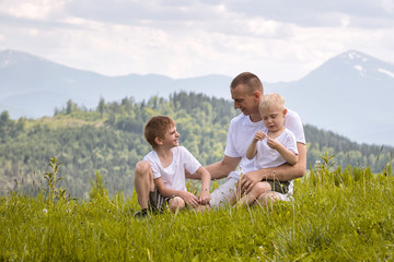 Fototapeta premium Happy father with his two young sons sitting on the grass on a background of green forest, mountains and sky with clouds. Friendship concept.