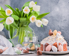 Obraz na płótnie Canvas Bouquet of white tulips in a vase on a gray background. Shokloy eggs of different colors and Easter cake with cream, as a symbol of the Easter holiday. Spring card with flowers and sweets. Copy space.
