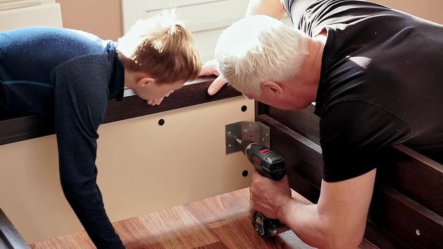 Grandfather and grandson are repairing furniture. Grandfather unscrews the screw with an electric screwdriver. Grandson helps him, raises from the floor the screw.