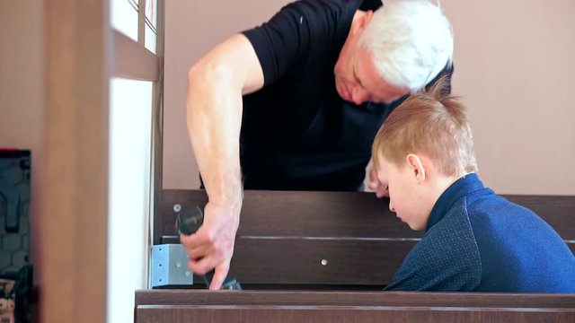 Grandfather and grandson are repairing furniture. Grandpa parses bed electric screwdriver, grandson sitting on the floor nearby and watching.