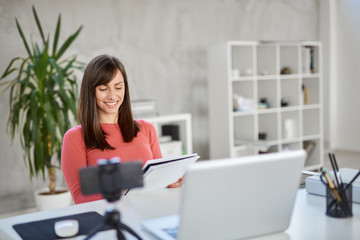 Charming Caucasian businesswoman sitting in modern office and taking notes in notebook. In front of her laptop.