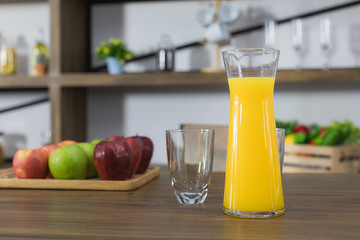 Close up image of a jug of freshly squeezed orange juice and drinking glass on the dark wooden table top,   with a blur green and red apple and kitchen room background
