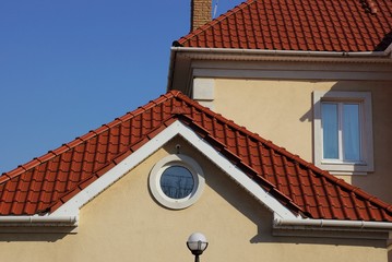 part of a brown house with a round window in the attic and a red tiled roof against the blue sky