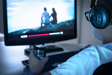 Guy watching movie or series online from streaming service and drinking can of soda. Internet on demand video player in computer. Tv show episodes marathon late at night. Young man with headphones.