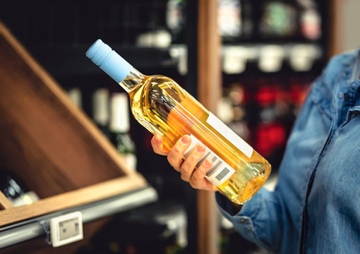 White Wine Bottle In Hand In Liquor Store. Customer Buying Alcohol. Woman Choosing The Right Bottle Of Chardonnay Or Riesling. Alcoholic Beverages Aisle And Shelf In Supermarket. Lady Shopping Drinks.