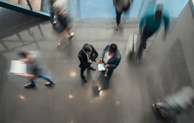 Two business people standing in the lobby of an office looking at a tablet while people are walking past in a blur - Powered by Adobe