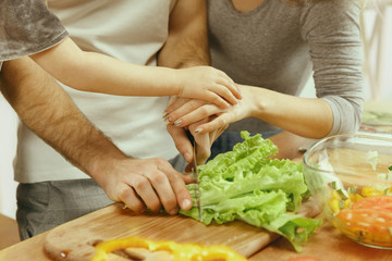 Cute little girl and her beautiful parents are cutting vegetables and smiling while making salad in kitchen at home. Family lifestyle concept. Hands close up