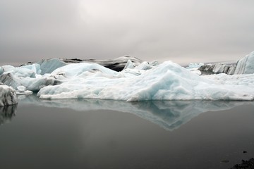 Reflections of crystal blue and white icerbergs in black dark water in dim gloomy light -...