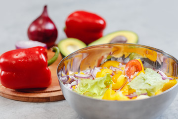 Bowl with sliced vegetables for fresh wholesome salad, in background are ingredients: avocado, pepper, onion, tomato