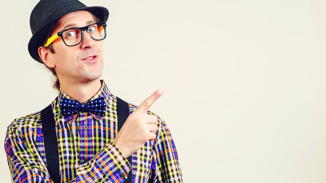 Male nerd in spectacles points with thumb on copy space for your advertising content. Funny nerd or geek on white background. Young funny man wears checkered shirt, suspender and hat.