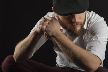 young handsome man in glasses with a beard and a mustache in a black cap and a white shirt conceived bent on his hands, a dark studio background 