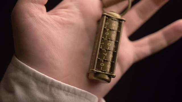 A man examines a copper metal cryptex with numbers, worn on a finger, through a leather strap loop. Then squeezes the cryptex in his hand. Close-up.