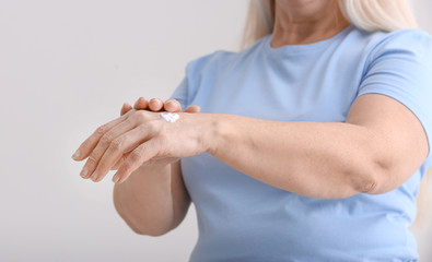 Mature woman applying hand cream on grey background