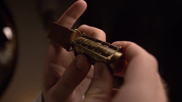 A man holds a copper cryptex with numbers. He calculates the cryptex code with his hand. At the end of the cryptex, a leather strap is put on the man&rsquo;s finger. There is a dim light in room. Closeup.