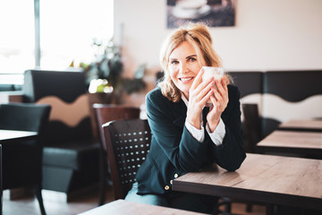 Attraktive,  ältere Frau sitzt im Cafe, hält einen Latte Macchiato in der Hand und lächelt...